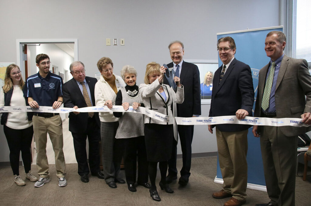 Members of the SMWC and the Terre Haute communities hold the ribbon and President Dottie L. King cuts it with large scissors.