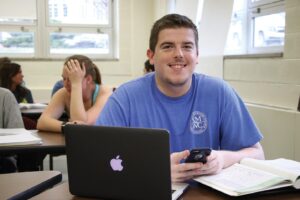 Austin Sievers sitting in classroom smiling, with books and laptop open
