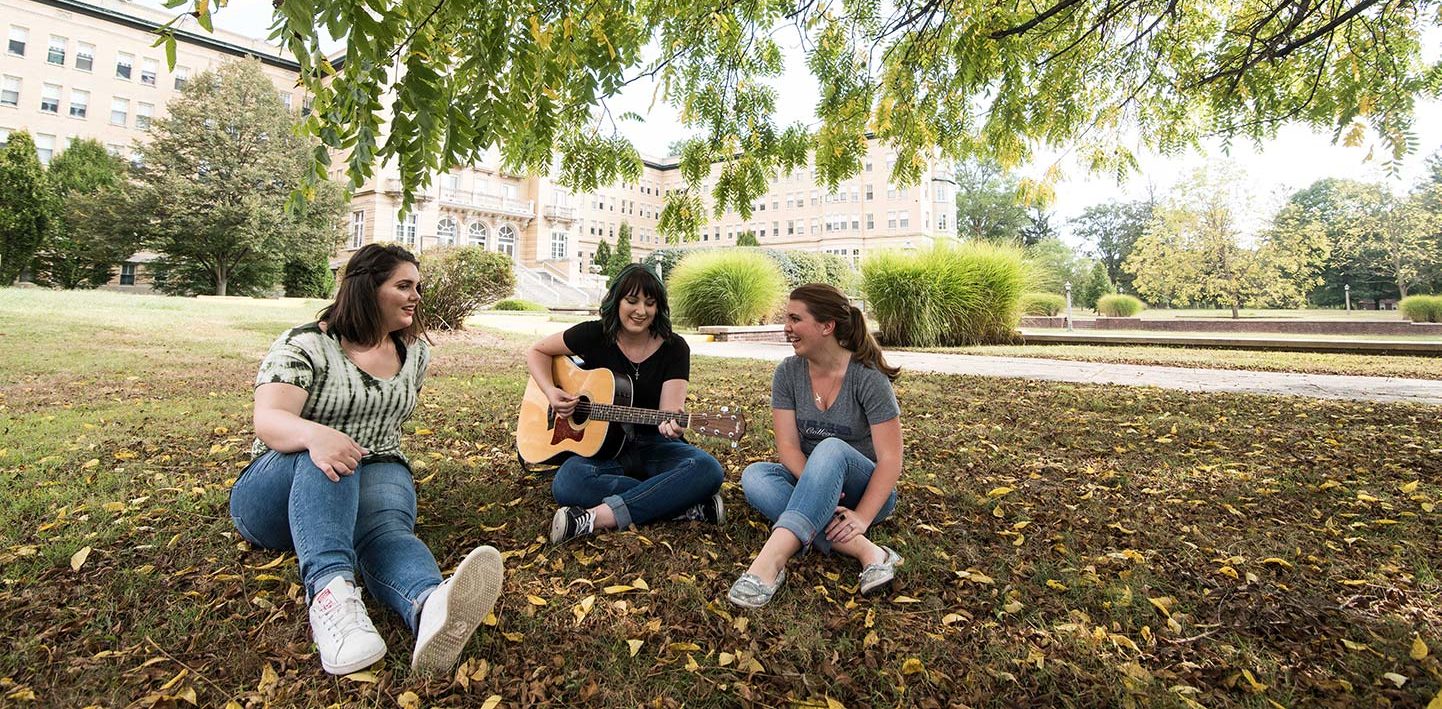Group of students sitting in the lawn in front of Le Fer Hall, with the woman in the middle playing a guitar