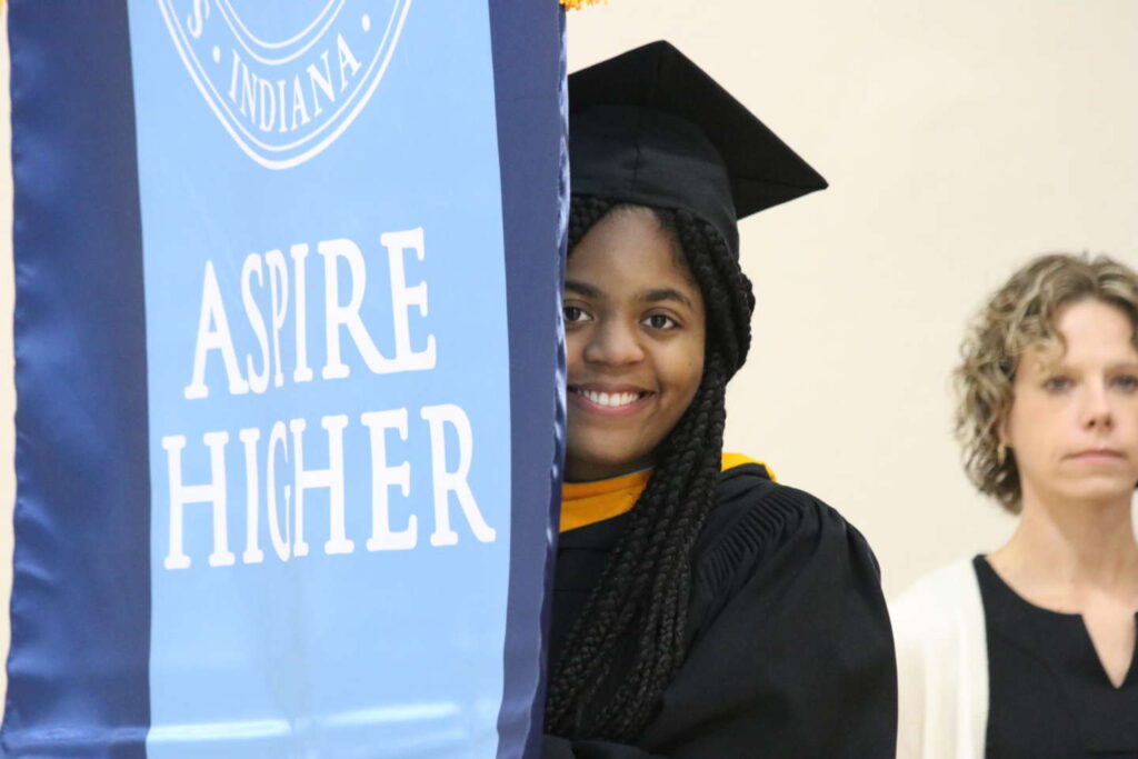 Graduate stands behind Aspire Higher banner at commencement.