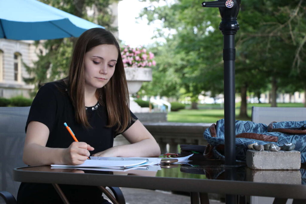 Student sits at a table outside making notes on a paper.