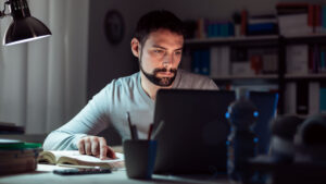 Young man sitting at desk, using a laptop, working late at night, he is studying a book and using a laptop