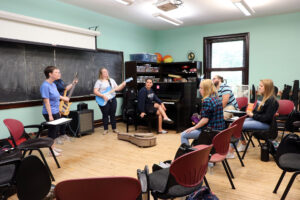 A music class with two people playing guitars, a student singing, and four students listening