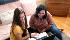 Two students sitting together on the floor looking at a laptop in a dorm room