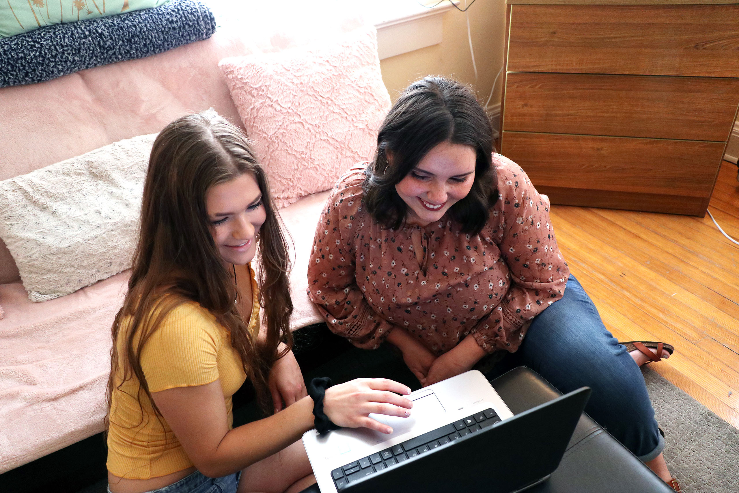 Two students sitting together on the floor looking at a laptop in a dorm room