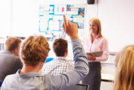 High school students with their hands up while a teacher is in the front of class teaching.