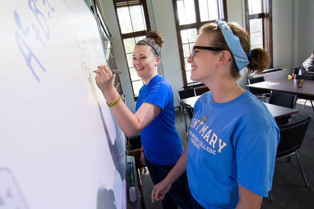 Two female students standing at a dry erase board working together on homework.