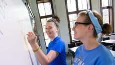 Two female students standing at a dry erase board working together on homework.