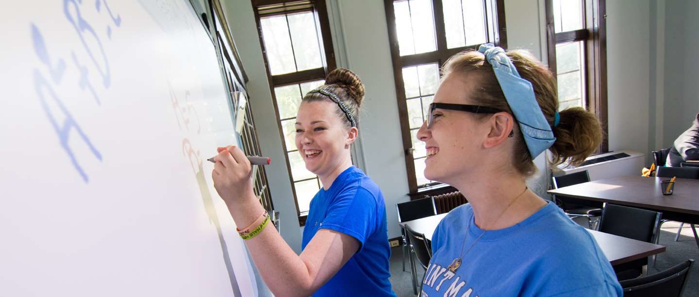 Two female students standing at a dry erase board working together on homework.