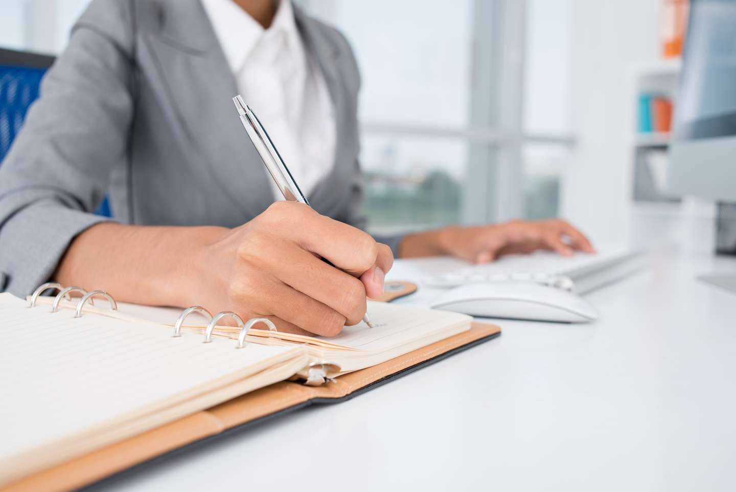 The hands of a female administrator working at her computer and writing in a binder.