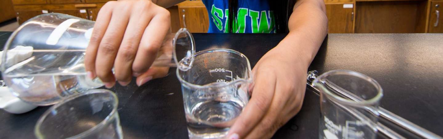 Someone's hands on a chemistry beaker pouring liquid into a glass.