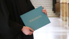 The hands of someone in a graduation gown holding a degree jacket that says Saint Mary-of-the-Woods College.