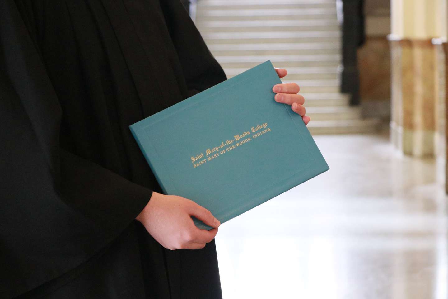 The hands of someone in a graduation gown holding a degree jacket that says Saint Mary-of-the-Woods College.