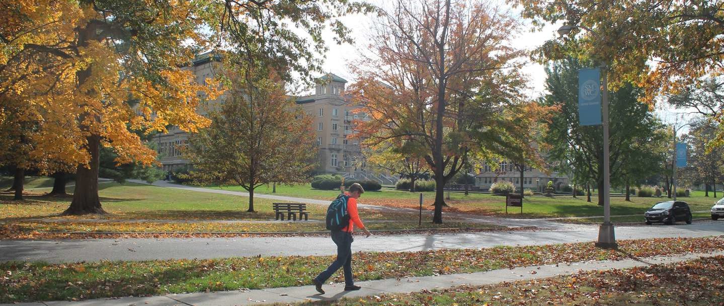 A student walks across campus with the fall leaves and Le Fer Hall in the background.