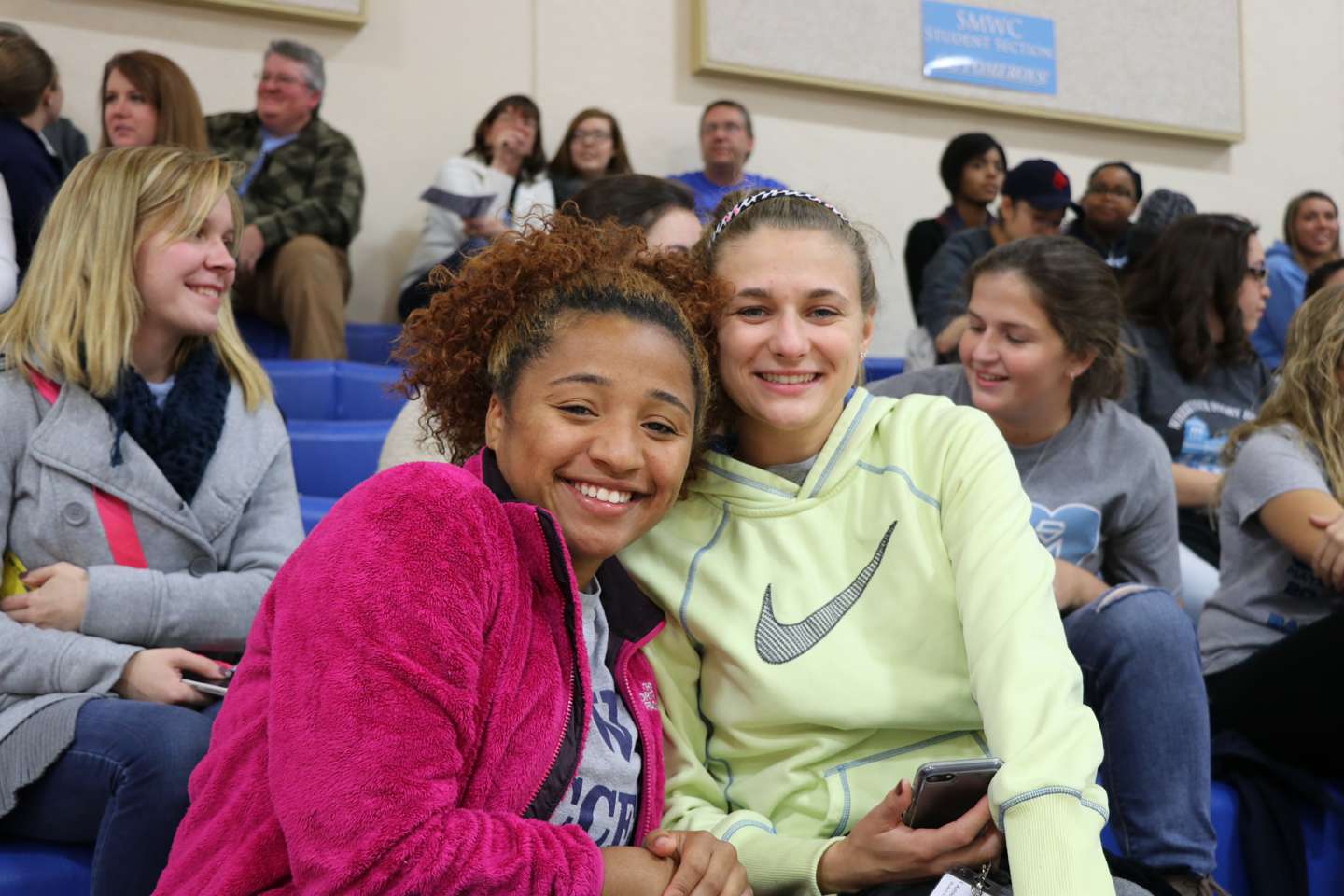Two friends sitting in the bleachers, holding a phone and smiling.